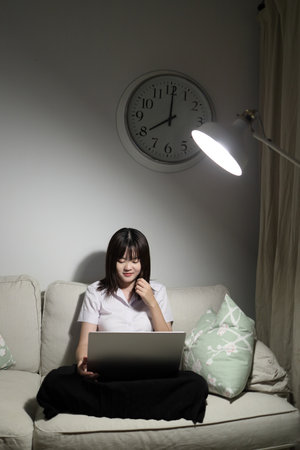 A young female student in a Thai university uniform sits on a sofa in the living room, studying online with a laptop. International Students' Day, World Students' Day. Gen Z, E-learningの写真素材