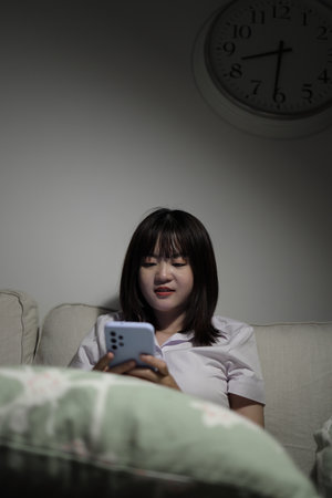A young female student in a Thai university uniform sits on a sofa in the living room, studying online with an application on mobile phone. International Students' Day, Gen Z, E-learningの写真素材