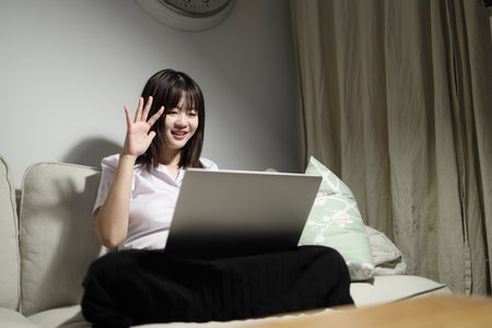 A young female student in a Thai university uniform sits on a sofa in the living room, studying online with a laptop. International Students' Day, World Students' Day. Gen Z, E-learningの写真素材