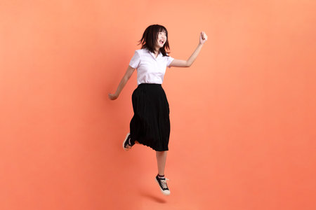 Cheerful female student in Thai university uniform with gesture of Jump isolated on orange background. International Students' Day, World Students' Day. Gen Zの写真素材