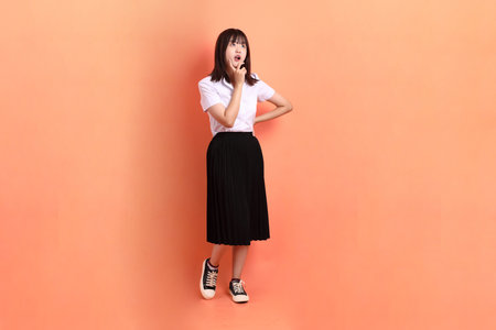 Cheerful female student in Thai university uniform with gesture of  thinking isolated on orange background. International Students' Day, World Students' Day. Gen Zの写真素材
