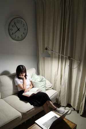 A young female student in a Thai university uniform sits on a sofa in the living room, reading a book. International Students' Day, World Students' Day. Gen Zの写真素材