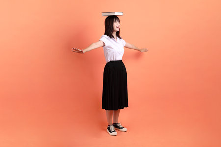 Cheerful female student in Thai university uniform with gesture of holding a textbook isolated on orange background. International Students' Day, World Students' Day. Gen Zの写真素材