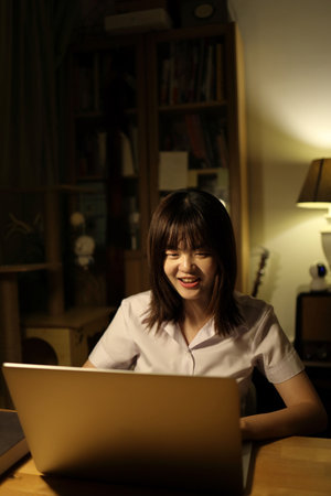 A young female student in a Thai university uniform sits at a desk in study, studying online with a laptop. International Students' Day, World Students' Day. Gen Z, E-learningの写真素材