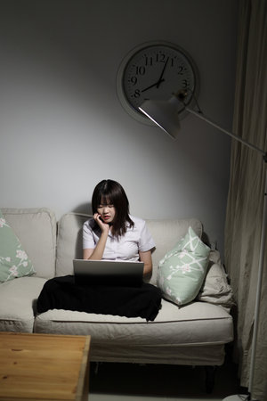 A young female student in a Thai university uniform sits on a sofa in the living room, studying online with a laptop. International Students' Day, World Students' Day. Gen Z, E-learningの写真素材