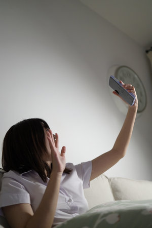 A young female student in a Thai university uniform sits on a sofa in the living room, studying online with an application on mobile phone. International Students' Day, Gen Z, E-learningの写真素材