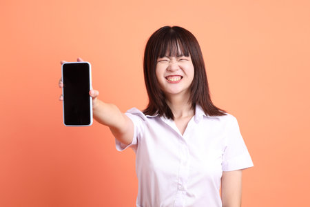 Cheerful female student in Thai university uniform with gesture of Holding mobile phone isolated on orange background. International Students' Day, World Students' Day. Gen Zの写真素材