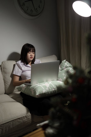 A young female student in a Thai university uniform sits on a sofa in the living room, studying online with a laptop. International Students' Day, World Students' Day. Gen Z, E-learningの写真素材