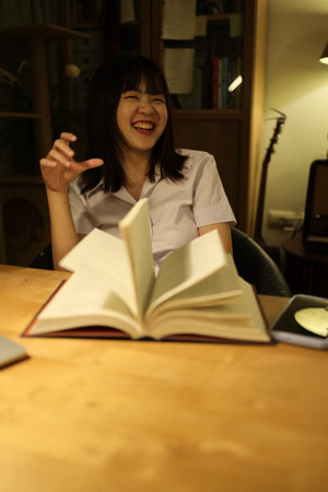 A young female student in a Thai university uniform sits at a desk in study, doing homework and...