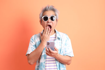 HAPPY SONGKRAN DAY. Asian tourist senior man in summer clothing with gesture of hand Holding mobile phone isolated on orange background. Songkran festival. Thai New Year's Day.の写真素材