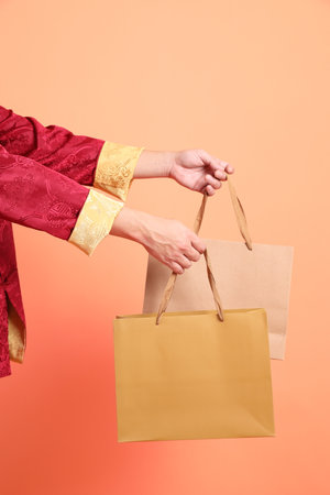 Happy Chinese new year. Hand of asian chinese senior man wearing red traditional clothing with gesture of hand holding gold paper shopping bags isolated on orange background.の写真素材