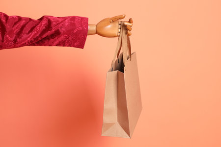 Happy Chinese new year. Wood art mannequin wearing red traditional clothing with gesture of hand holding brown paper shopping bag isolated on orange background.の写真素材