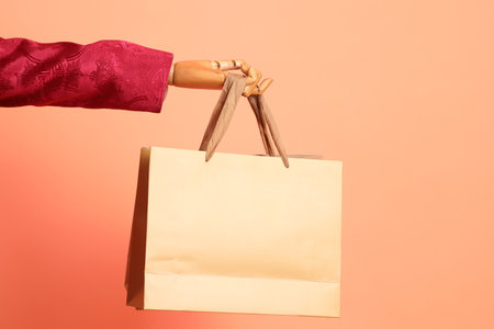 Happy Chinese new year. Wood art mannequin wearing red traditional clothing with gesture of hand holding brown paper shopping bags isolated on orange background.の写真素材