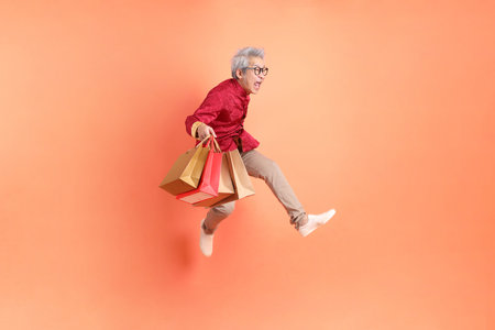Happy Chinese new year. Asian Chinese energetic senior man wearing red traditional clothing with gesture of hand holding paper shopping bags while jumping isolated on orange background.の写真素材