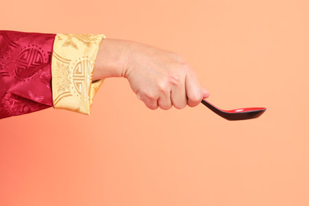 Happy Chinese new year. Hand of asian chinese senior man wearing red traditional clothing with gesture of hand holding spoon isolated on orange background.の写真素材