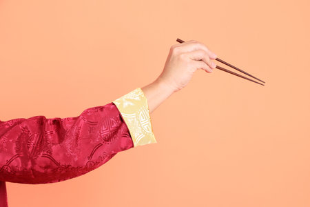 Happy Chinese new year. Hand of asian chinese senior man wearing red traditional cheongsam qipao or changshan dress with gesture of hand holding brown chopsticks isolated on orange background.の写真素材