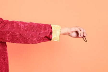 Happy Chinese new year. Hand of asian chinese senior man wearing red traditional cheongsam qipao or changshan dress with gesture of hand holding brown chopsticks isolated on orange background.の写真素材