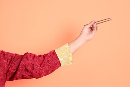 Happy Chinese new year. Hand of asian chinese senior man wearing red traditional cheongsam qipao or changshan dress with gesture of hand holding silver chopsticks isolated on orange background.の写真素材