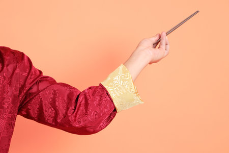 Happy Chinese new year. Hand of asian chinese senior man wearing red traditional cheongsam qipao or changshan dress with gesture of hand holding silver chopsticks isolated on orange background.の写真素材