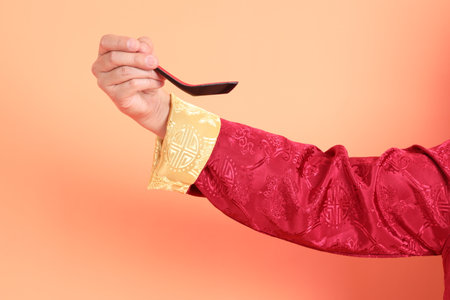 Happy Chinese new year. Hand of asian chinese senior man wearing red traditional clothing with gesture of hand holding spoon isolated on orange background.の写真素材