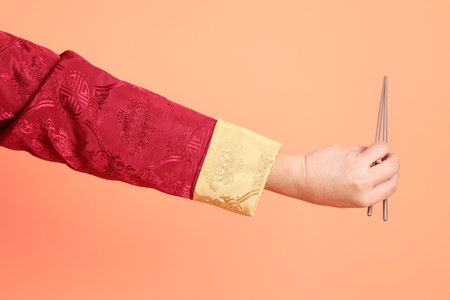 Happy Chinese new year. Hand of asian chinese senior man wearing red traditional cheongsam qipao or changshan dress with gesture of hand holding silver chopsticks isolated on orange background.の写真素材