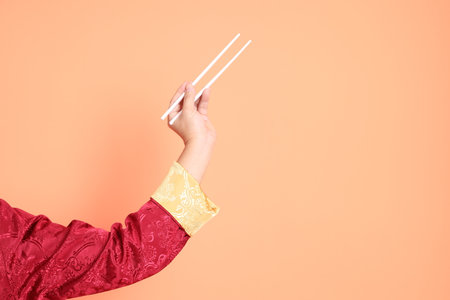 Happy Chinese new year. Hand of asian chinese senior man wearing red traditional cheongsam qipao or changshan dress with gesture of hand holding white chopsticks isolated on orange background.の写真素材