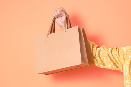 Happy Chinese new year. Hand of asian chinese senior man wearing golden traditional clothing with gesture of hand holding paper shopping bags isolated on orange background.の写真素材