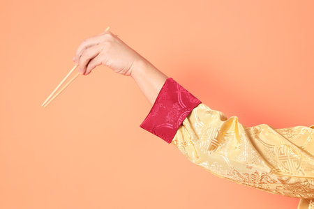 Happy Chinese new year. asian chinese senior man wearing golden traditional cheongsam qipao or changshan dress with gesture of hand holding wood chopsticks isolated on orange background.の写真素材