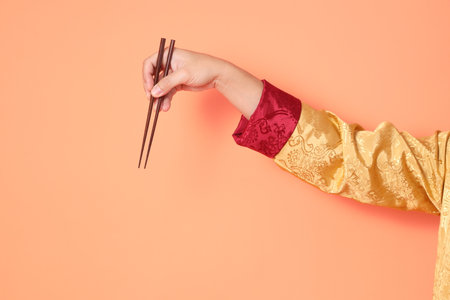 Happy Chinese new year. asian chinese senior man wearing golden traditional cheongsam qipao or changshan dress with gesture of hand holding brown chopsticks isolated on orange background.の写真素材