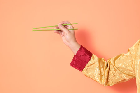 Happy Chinese new year. asian chinese senior man wearing golden traditional cheongsam qipao or changshan dress with gesture of hand holding green chopsticks isolated on orange background.の写真素材