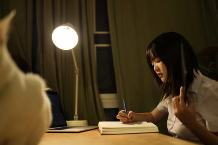 A young female student in a Thai university uniform sits at a desk in study, doing homework and study hard. International Students' Day, World Students' Day. Gen Z, E-learningの写真素材
