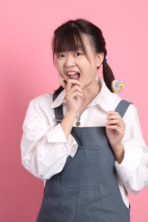 Cheerful lovely young asian woman in overalls casual clothes with gesture of holding heart shaped lollipop isolated on pink background. St Valentine's Day, Women's Day, Birthday, Sweet tooth conceptの写真素材