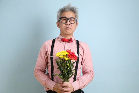 Asian senior man in black suspenders with red bow with gesture of holding a bouquet of flowers isolated on blue background. St Valentine's Day, Women's Day, Birthdayの写真素材