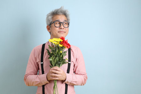 Asian senior man in black suspenders with red bow with gesture of holding a bouquet of flowers isolated on blue background. St Valentine's Day, Women's Day, Birthdayの写真素材