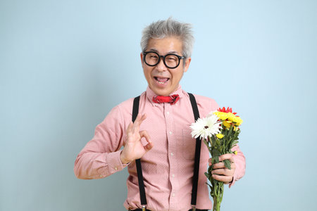 Asian senior man in black suspenders with red bow with gesture of holding a bouquet of flowers isolated on blue background. St Valentine's Day, Women's Day, Birthdayの写真素材
