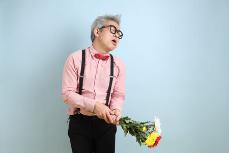 Asian senior man in black suspenders with red bow with gesture of holding a bouquet of flowers isolated on blue background. St Valentine's Day, Women's Day, Birthdayの写真素材