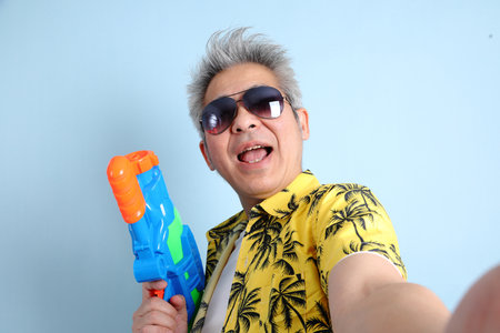 HAPPY SONGKRAN DAY. Asian energetic senior man in summer clothing and sunglasses with gesture of holding plastic water gun while take a selfie isolated on blue background. Songkran festival. Thai New Year's Day.の写真素材
