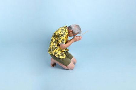 HAPPY SONGKRAN DAY. Asian tourist senior man in summer clothing and sunglasses with gesture of hand holding the incense sticks  isolated on blue background. Songkran festival. Thai New Year's Day.の写真素材