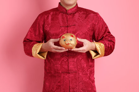 Happy Chinese new year. Asian Chinese energetic senior man wearing red traditional cheongsam qipao or changshan dress with gesture of holding Chinese piggy bank isolated on pink background.の写真素材