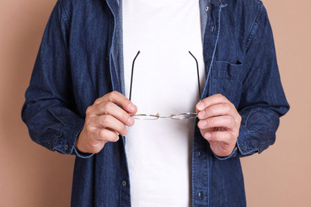 Asian manâ€™s hand holding a pair of glasses isolated on brown background, studio shot.