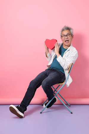Asian senior man in casual clothing with gesture of siting on the chair while holding holding red heart-shaped isolated on pink background. St Valentine's Dayの写真素材