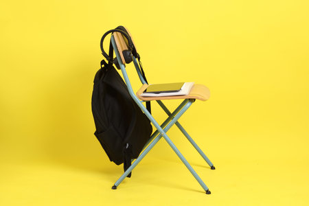 Back to school concept with school books, textbooks, backpack and stationery supplies on classroom chair isolated on yellow background. Studying abroad, Gen Z, E-learning.の写真素材