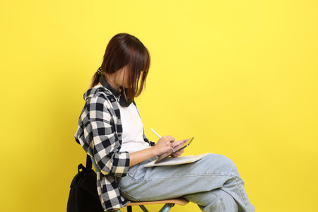 Asian college female student wearing plaid shirt carry school bag and book, gesture of sitting and Holding tablet. Studying abroad, Gen Z, E-learning.の写真素材