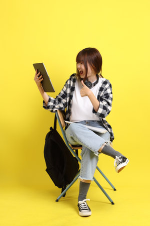 Asian college female student wearing plaid shirt carry school bag and book, gesture of sitting and Holding tablet. Studying abroad, Gen Z, E-learning.の写真素材