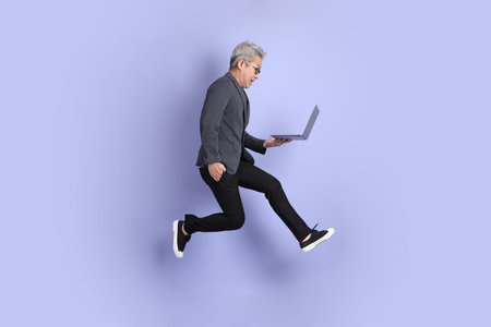 50s asian senior Businessman with glasses dressed in formal outfit with gesture of Holding laptop while jumping isolated on purple background.の写真素材