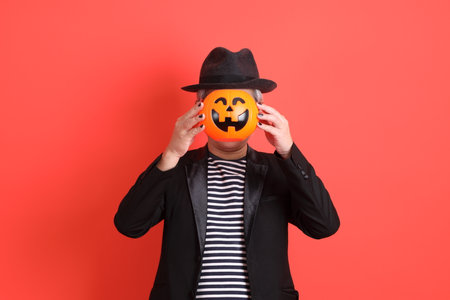 Asian senior man in Halloween costume with gesture of standing while hand holding Jack-o-Lantern Pumpkin isolated on orange background.の写真素材