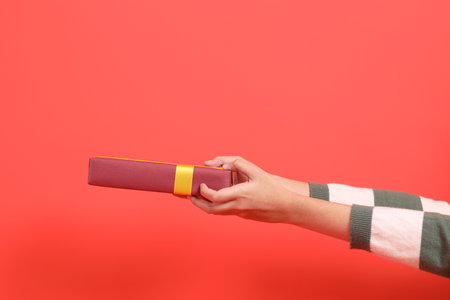 A close-up of a hand wearing a green stripe pattern sweater holding up a gift or present box, isolated against a red background. Merry Christmas.の写真素材
