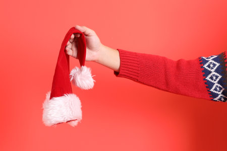 A close-up of a hand wearing a red cardigan holding up Santa Hat, isolated against a red background. Merry Christmas.の写真素材