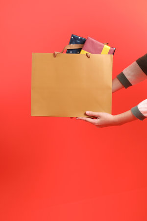 A close-up of a hand wearing a green stripe pattern sweater holding up a shopping bag, isolated against a red background. Merry Christmas.の写真素材