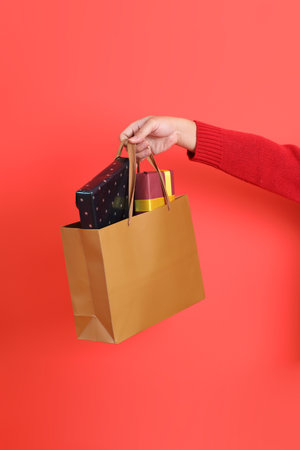 A close-up of a hand wearing a red cardigan holding up shopping bag, isolated against a red background. Merry Christmas.の写真素材